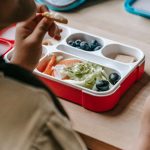 Crop faceless teenage sitting at wooden table with lunch box full of healthy food